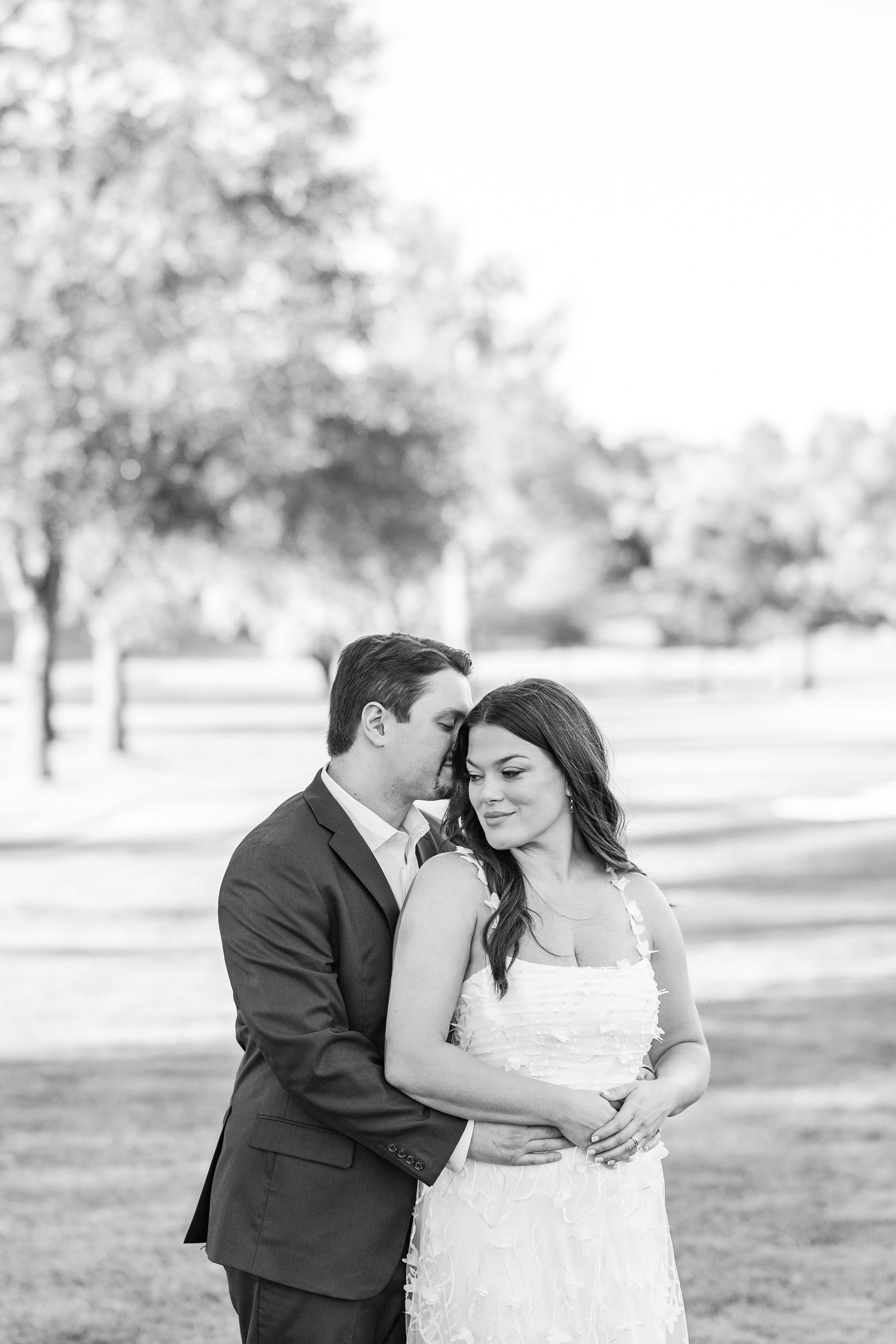 A romantic black and white portrait of a couple embracing outdoors in formal wedding attire on a sunny day.