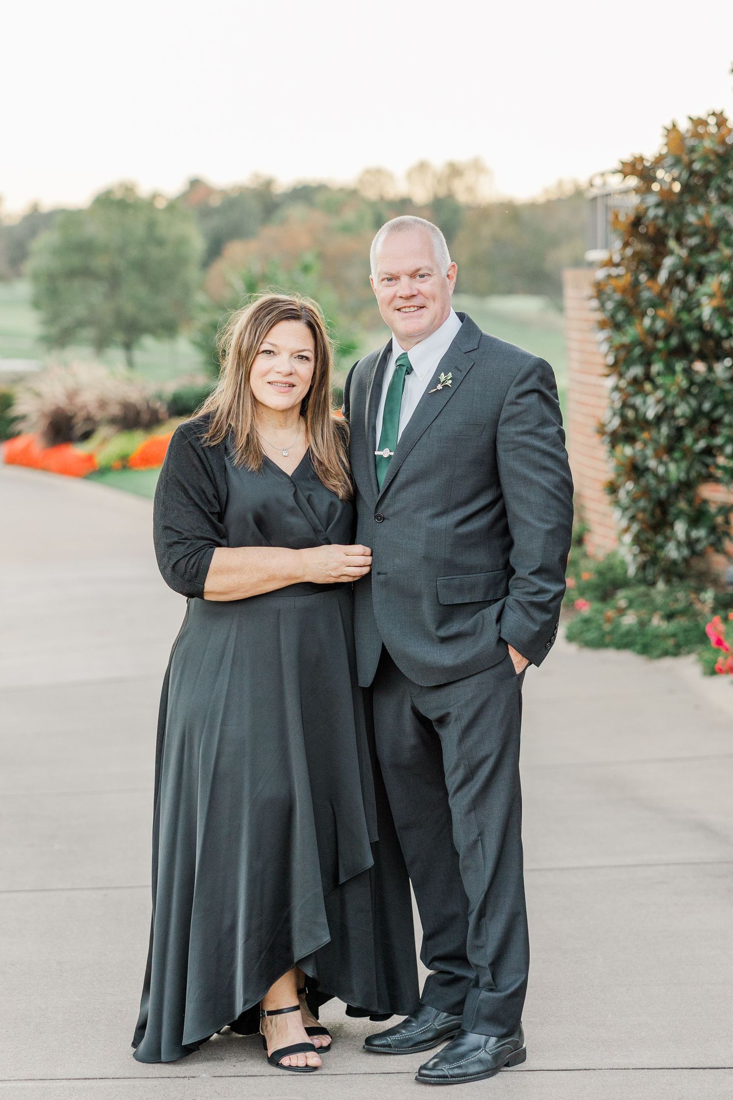 A couple poses together outdoors in formal attire with black dress and suit against a natural background.