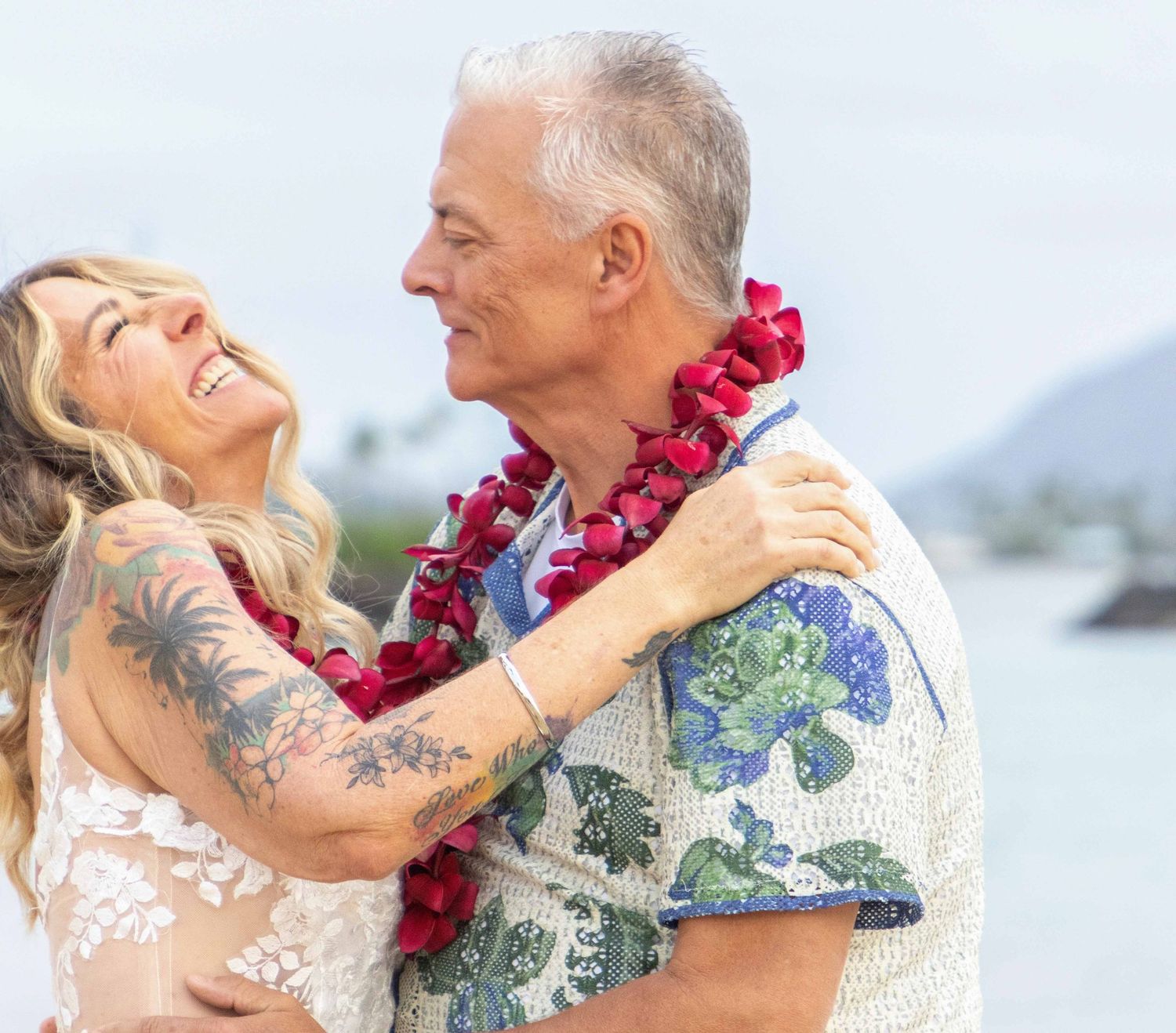A couple wearing Hawaiian leis and tropical shirts embrace joyfully on a sunny beach.