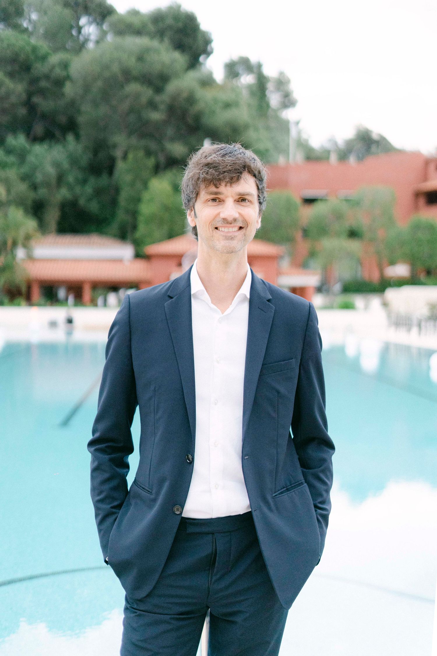 A person in a navy suit and white shirt stands with hands in pockets by a swimming pool with trees in the background.