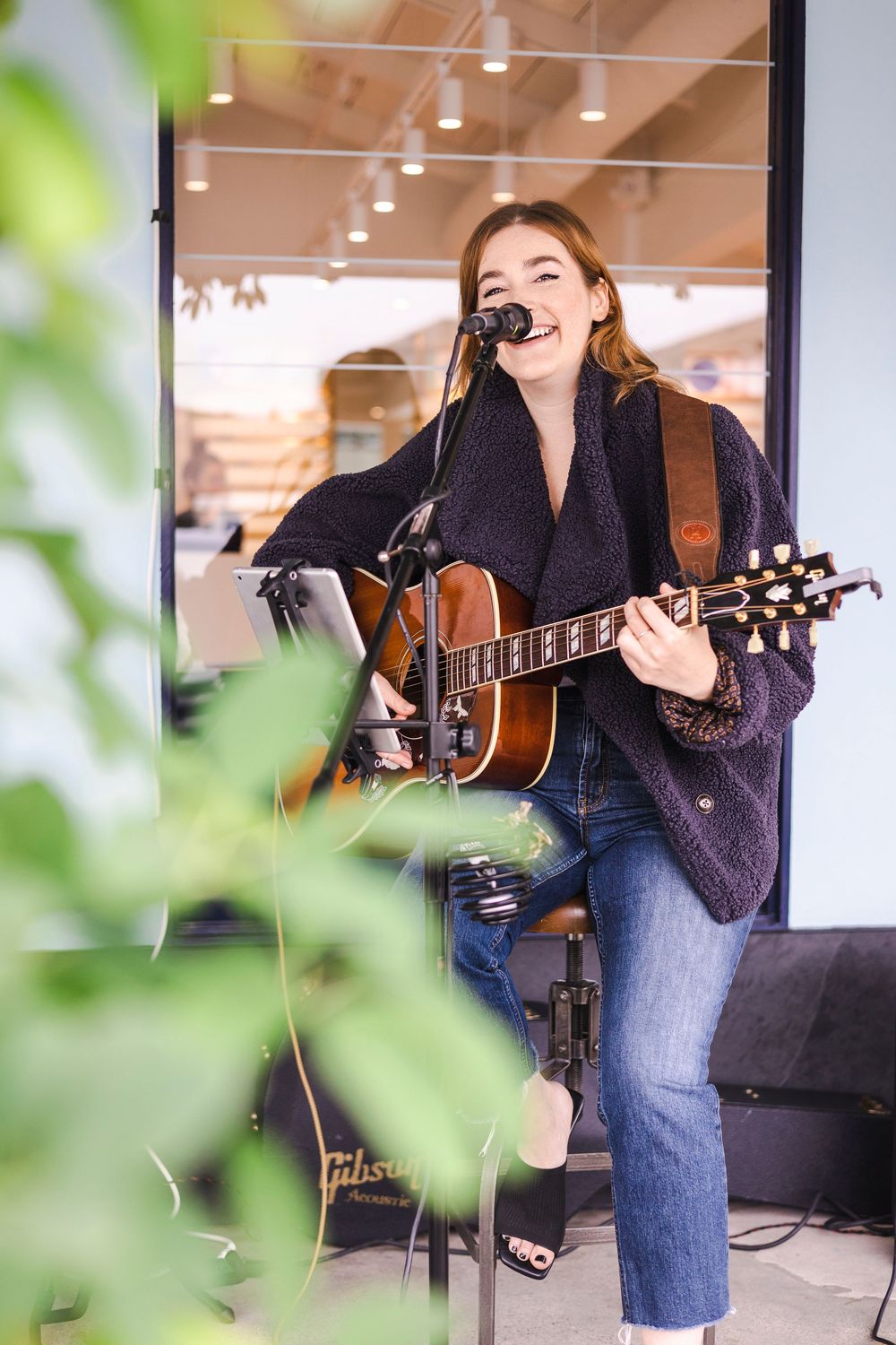 Musician performing with an acoustic guitar in a casual indoor venue with green foliage in foreground.