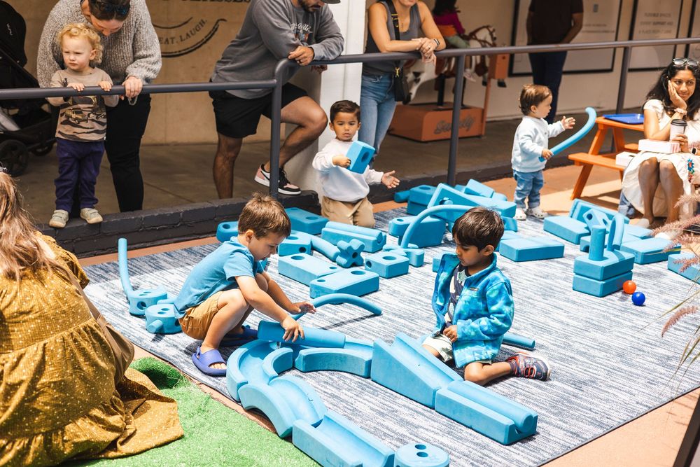Children in blue outfits sit on blankets during an indoor activity session with colorful building blocks and toys.