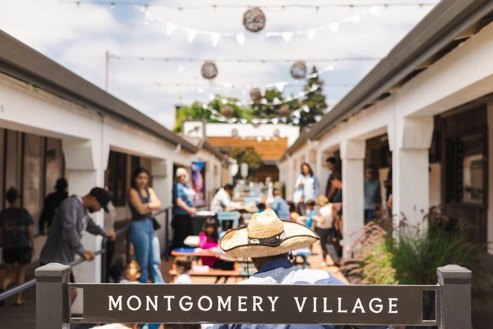 Shoppers browse outdoor stalls and walkways at Montgomery Village shopping center with hanging lights and storefronts.