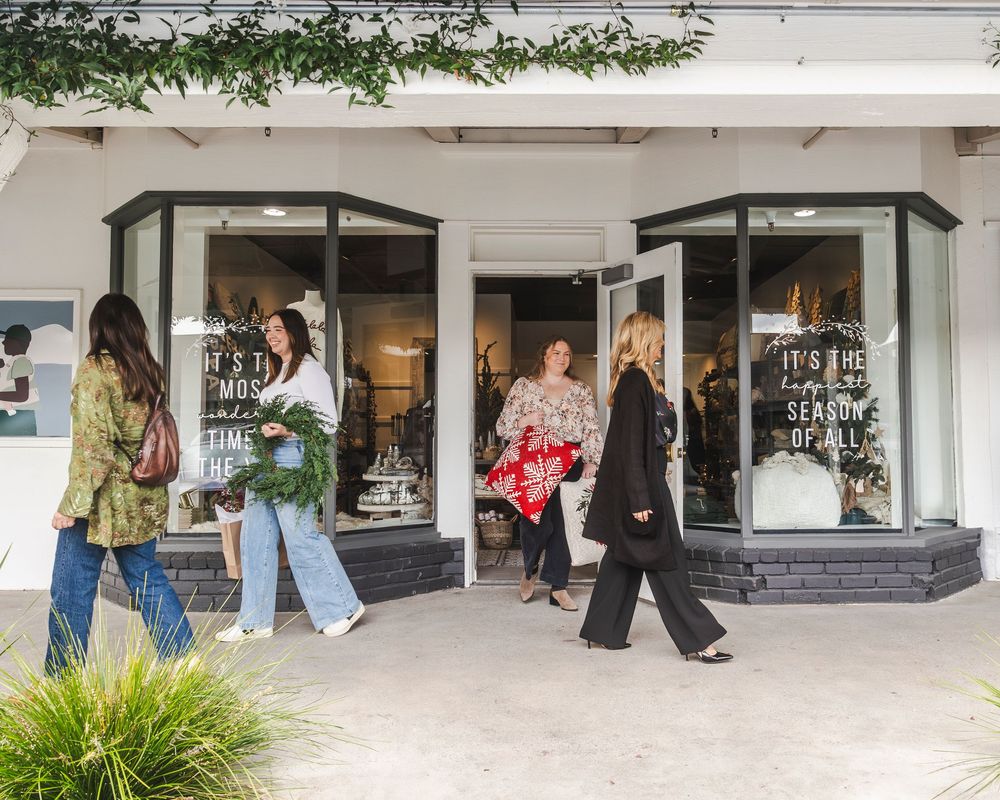 Customers browse retail storefronts along a modern shopping center walkway with white walls and tropical plants.