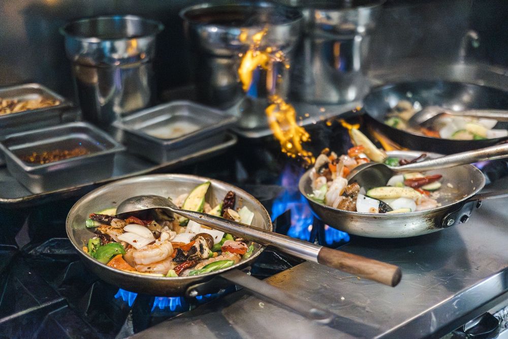 Professional chefs prepare dishes in a busy restaurant kitchen with flames rising from stovetops and multiple plates in progress.