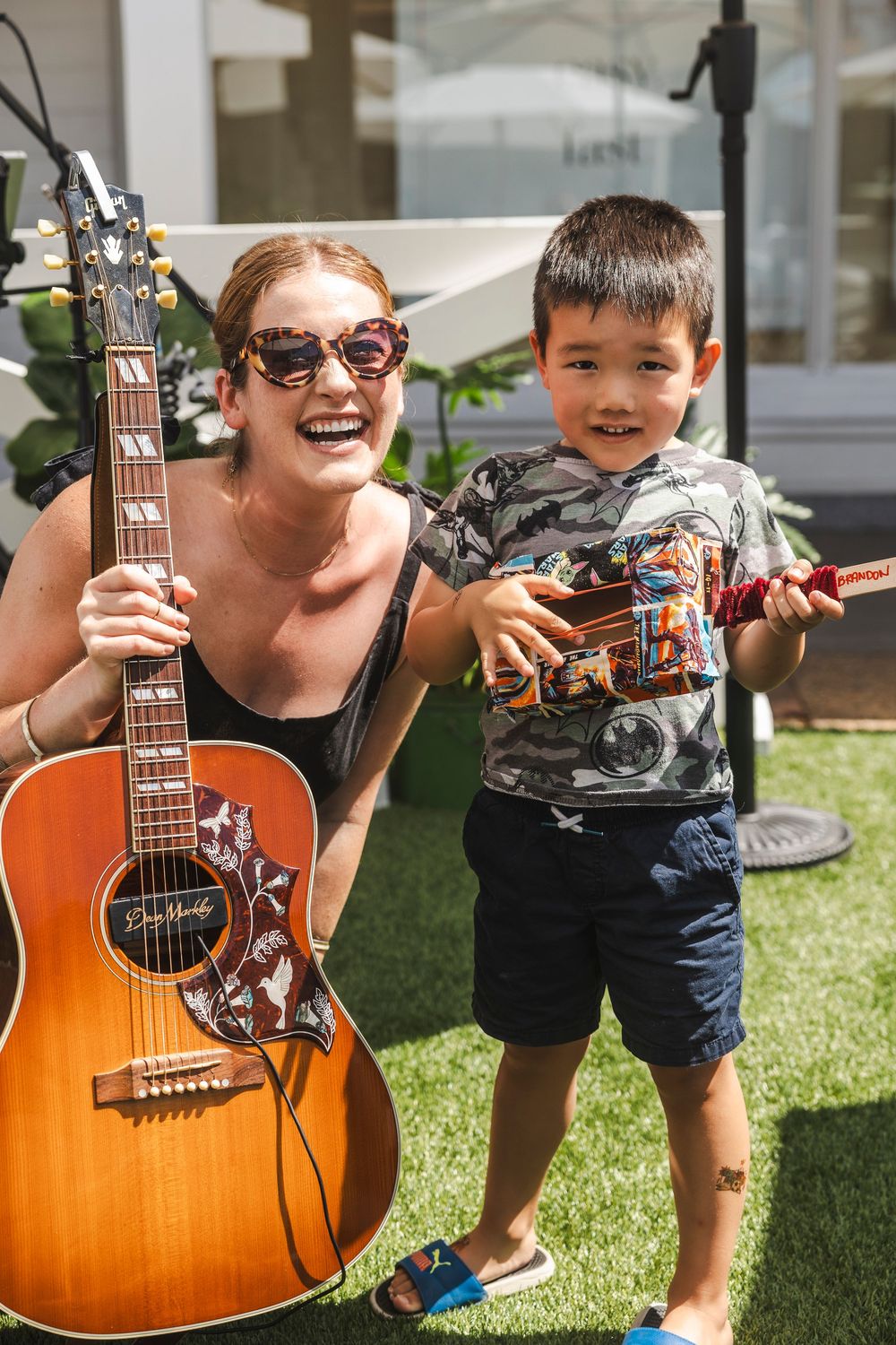 A person holding an acoustic guitar outdoors on grass while a small child plays nearby.