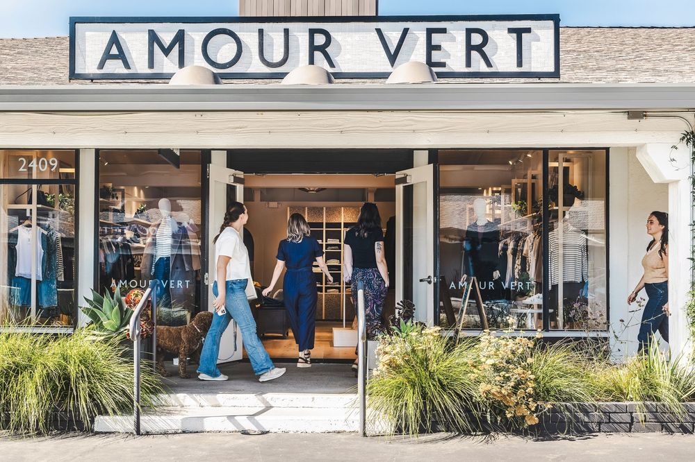 Storefront of Amour Vert boutique with window displays, ornamental grasses, and customers browsing clothing.