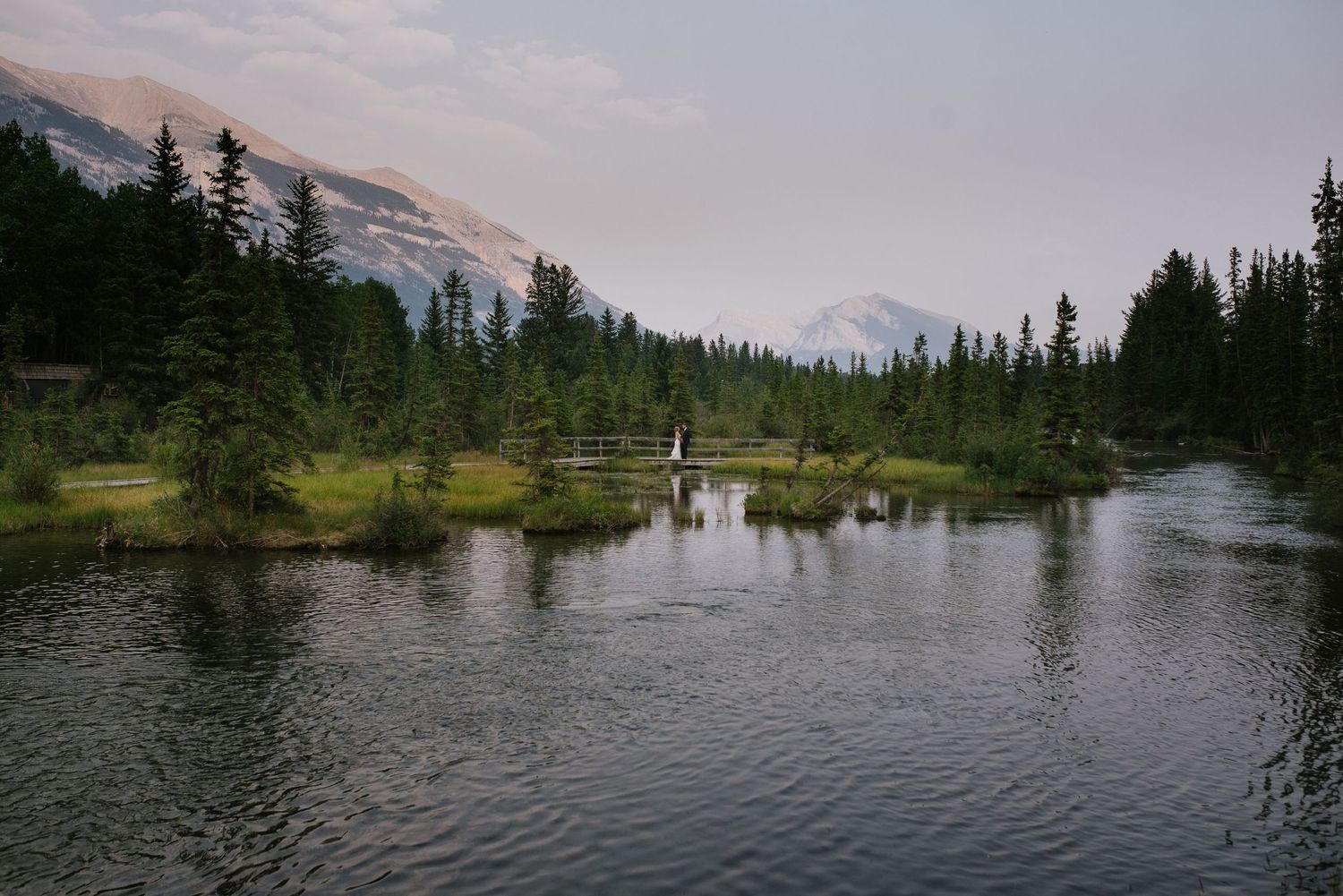 A policemen's creek in Canmore surrounded by  trees reflects the sky in a tranquil wilderness setting with a married couple