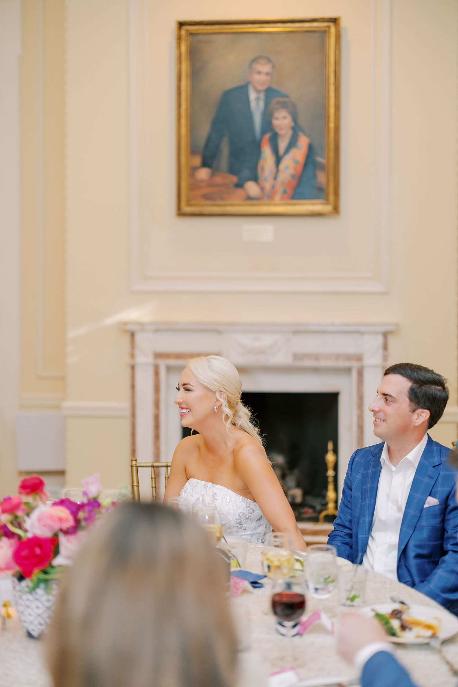 Couple seated at elegant wedding reception table with pink flowers and ornate fireplace in background.