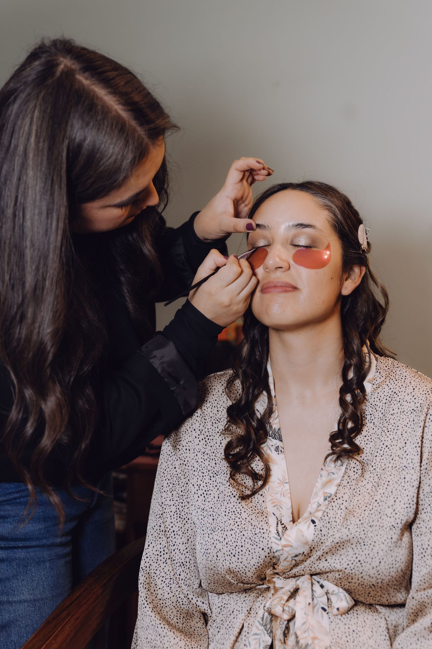 Makeup artist applies eye makeup to bride with pink eye patches during wedding preparation.