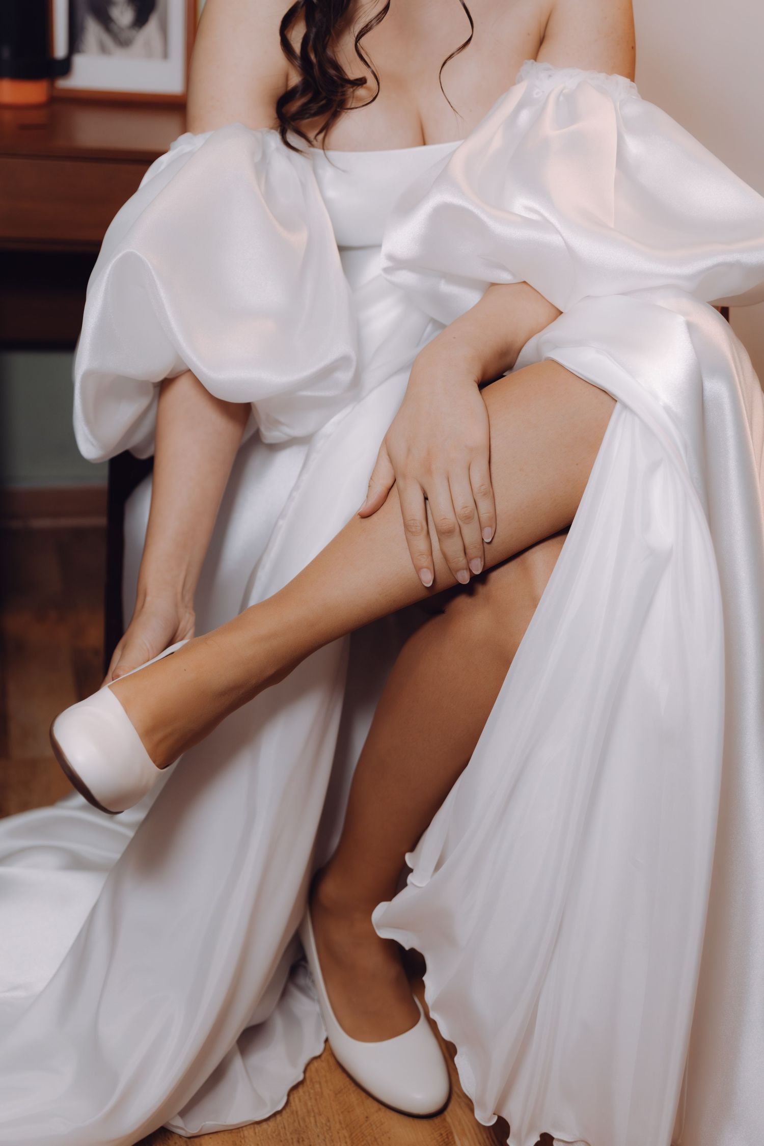 Bride in white satin gown with puff sleeves putting on white bridal shoes.