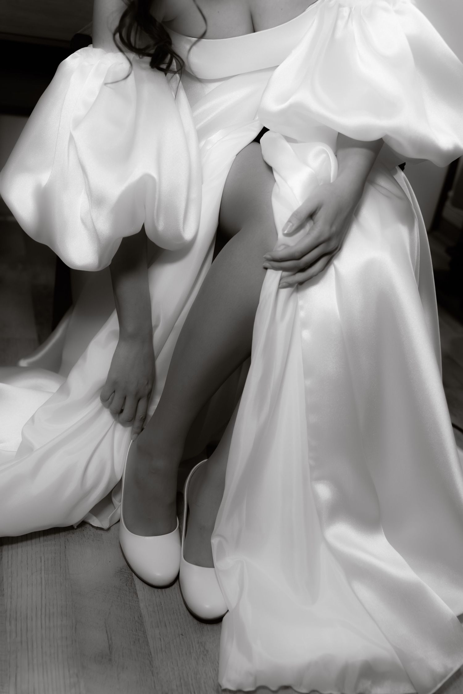 Black and white photo of bride in satin wedding dress with puff sleeves adjusting shoes.