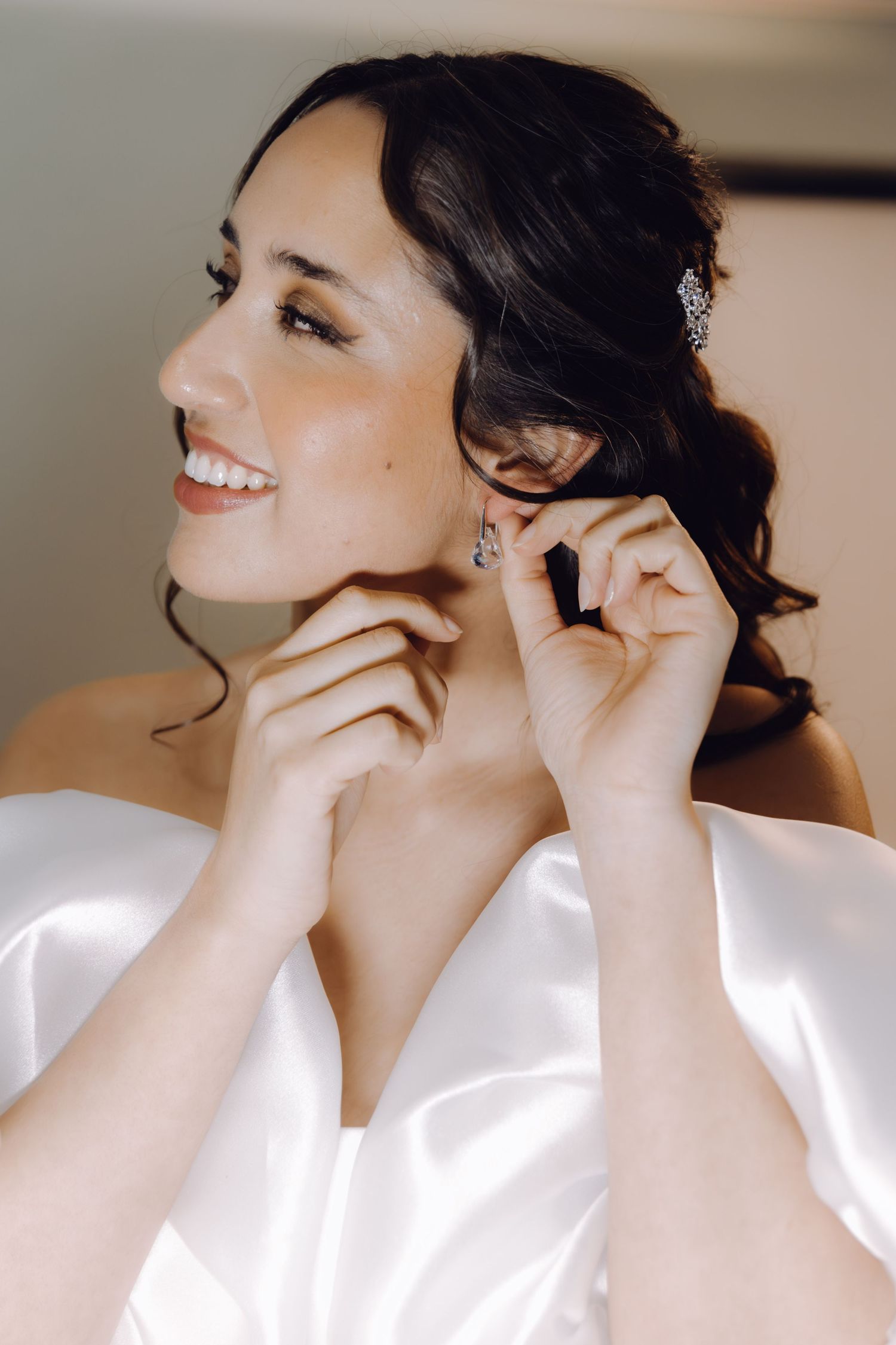 Smiling bride in white satin dress putting on crystal drop earrings on wedding day.