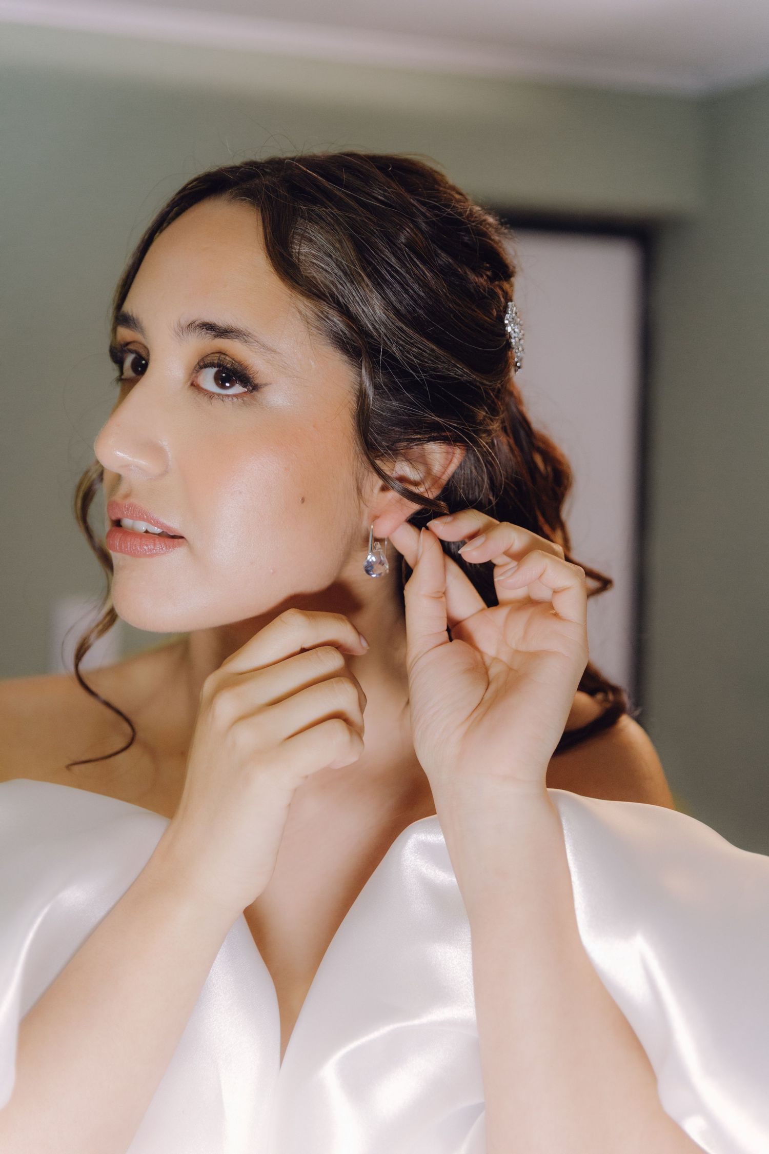 Beautiful bride in off-shoulder white gown having earrings fastened by bridesmaid.