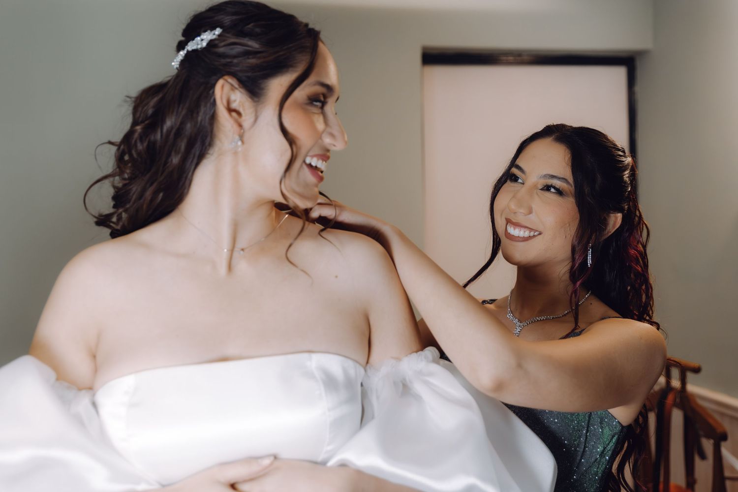 Bridesmaid in teal dress helping bride in white satin gown get ready for wedding.