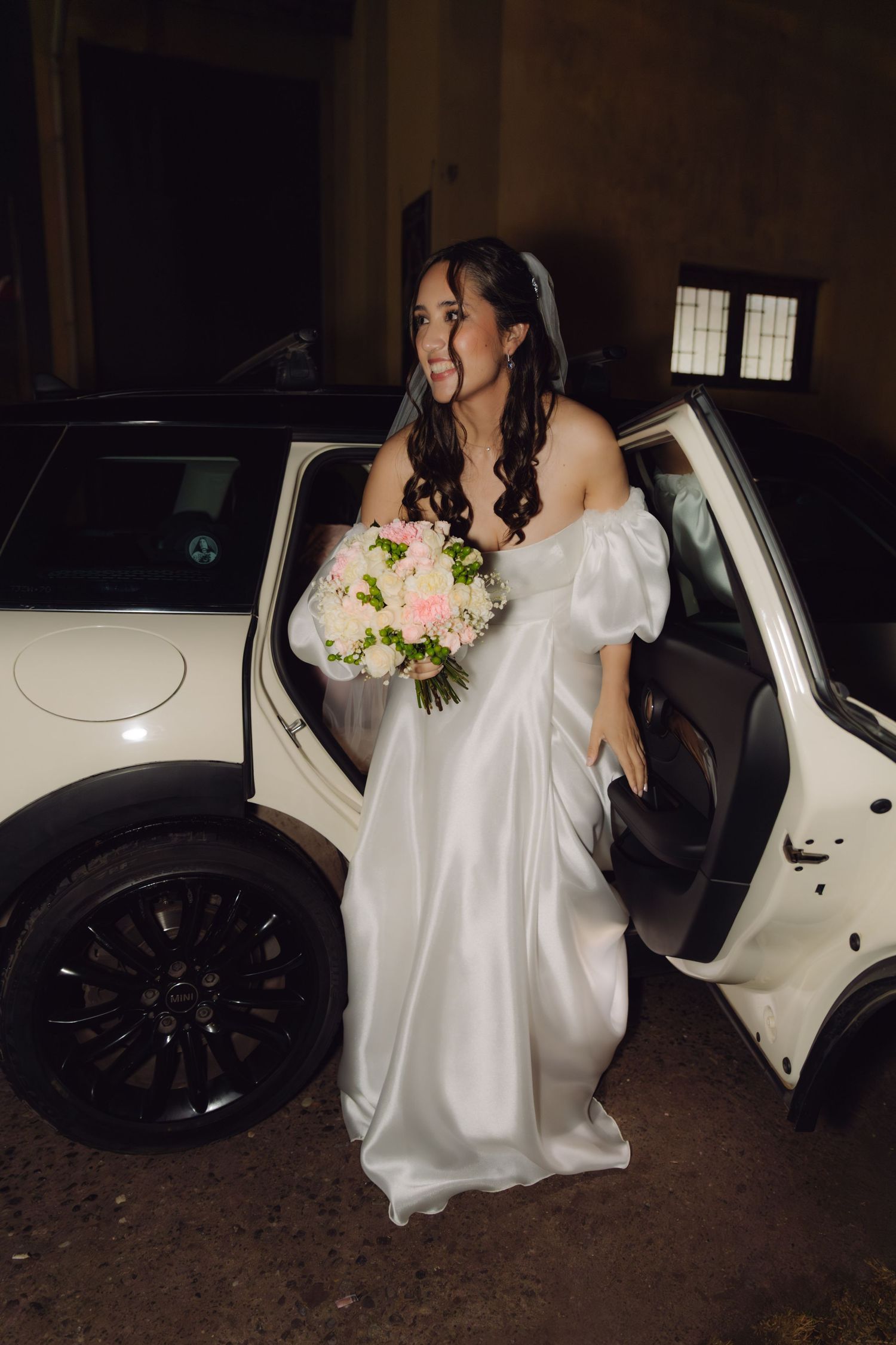 Excited bride in white satin puff sleeve dress stepping out of white Mini Cooper.