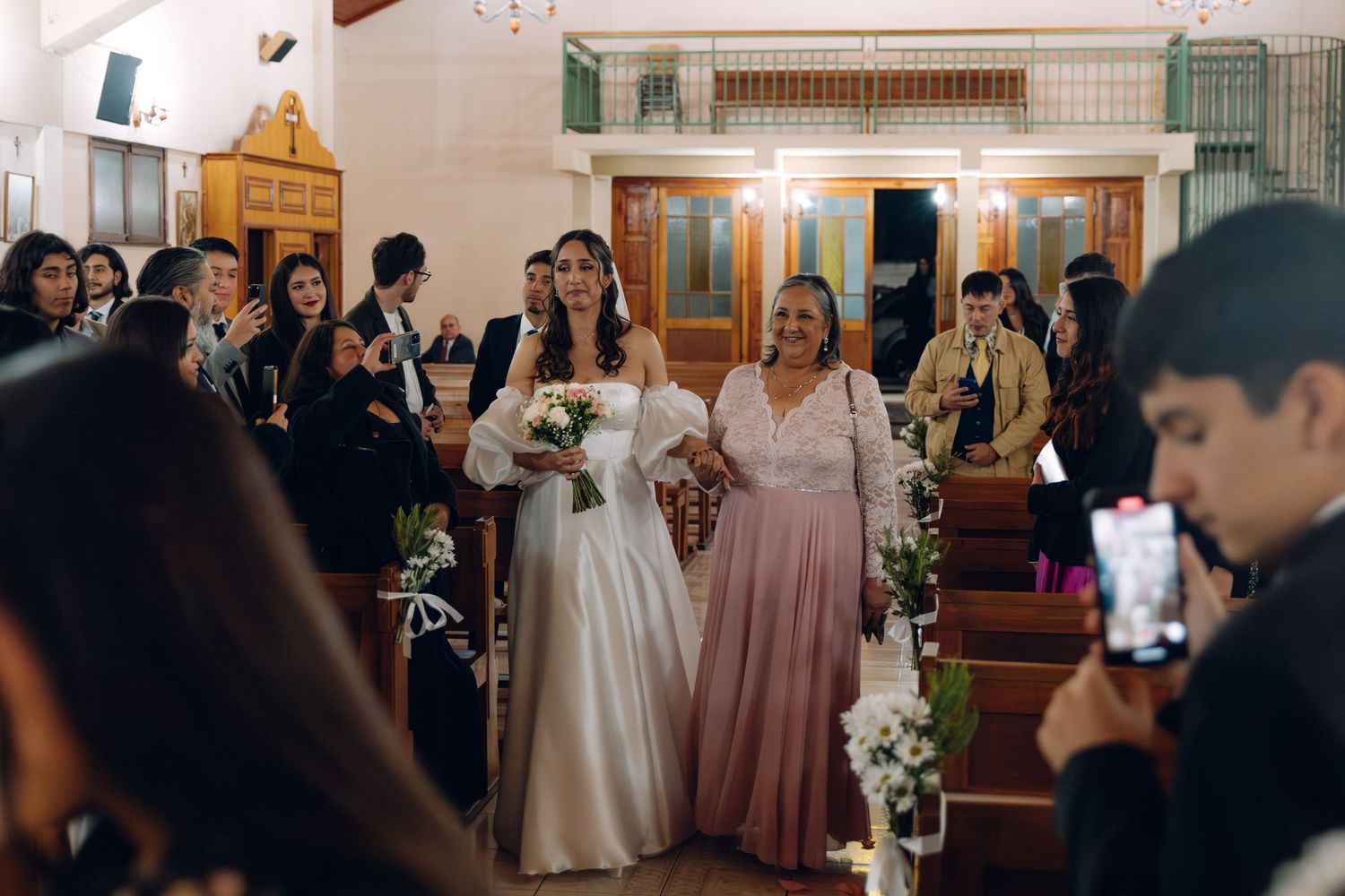 Bride walking down church aisle with mother in pink dress at wedding ceremony.