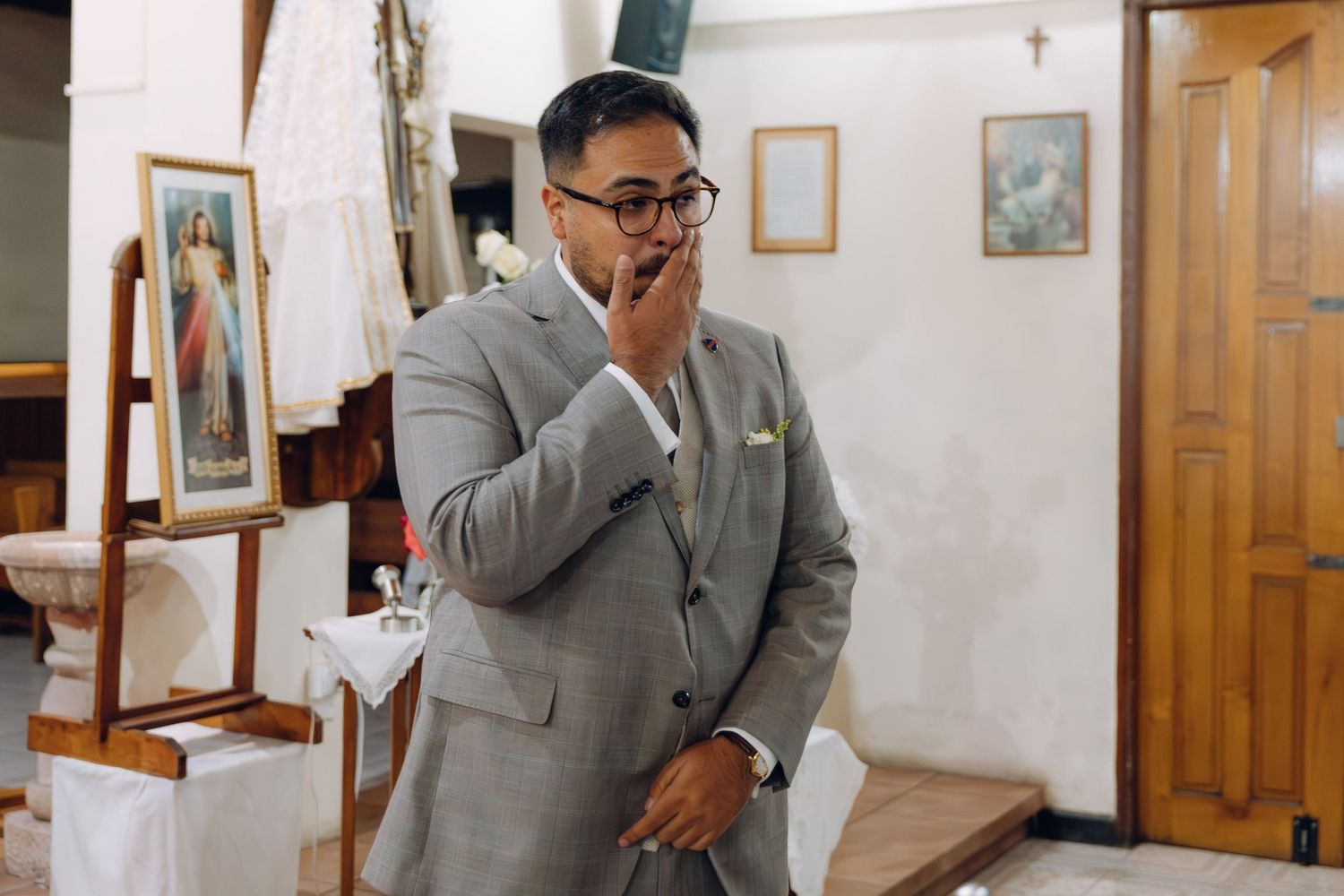 Emotional groom in gray suit covering his mouth in surprise inside a decorated Catholic church.