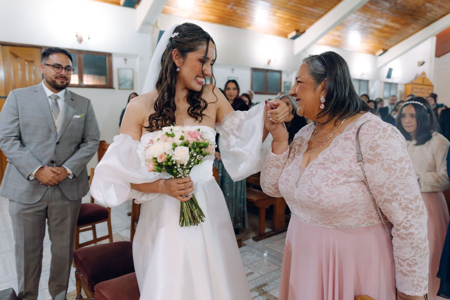 Bride in white gown with bouquet laughing with older woman in pink dress at wedding ceremony.
