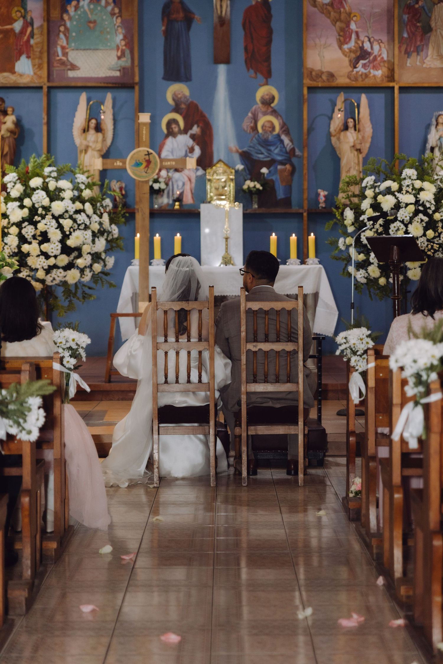 Wedding couple seated at altar in beautifully decorated Catholic church with floral arrangements.