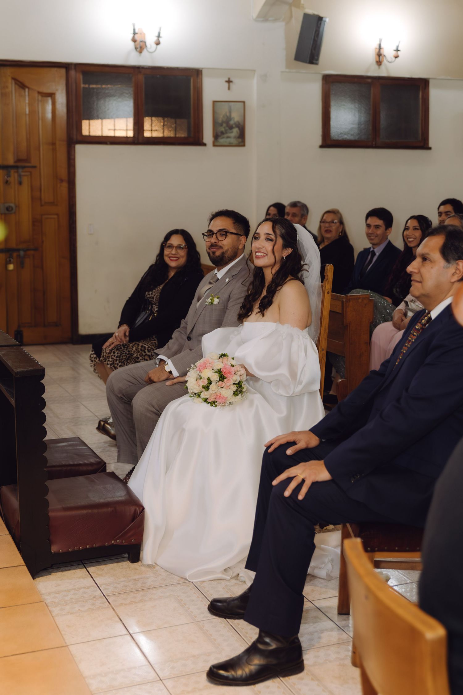 Smiling bride holding pink bouquet seated beside groom during church wedding ceremony.