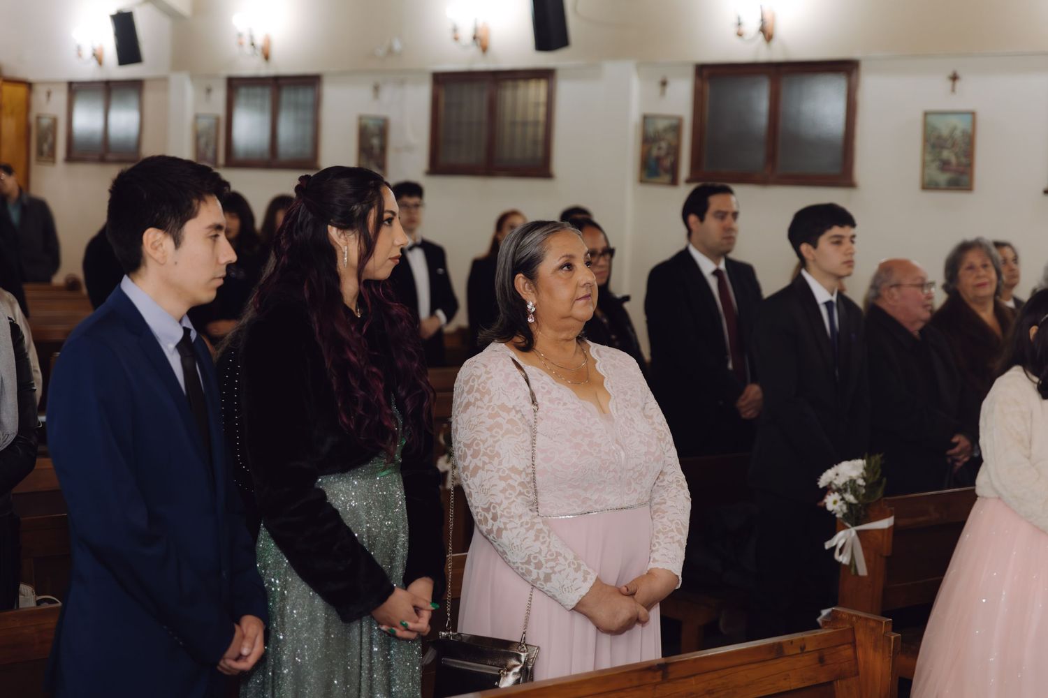 Wedding guests stand in church pews during a ceremony, including a woman in a pink dress in the foreground.
