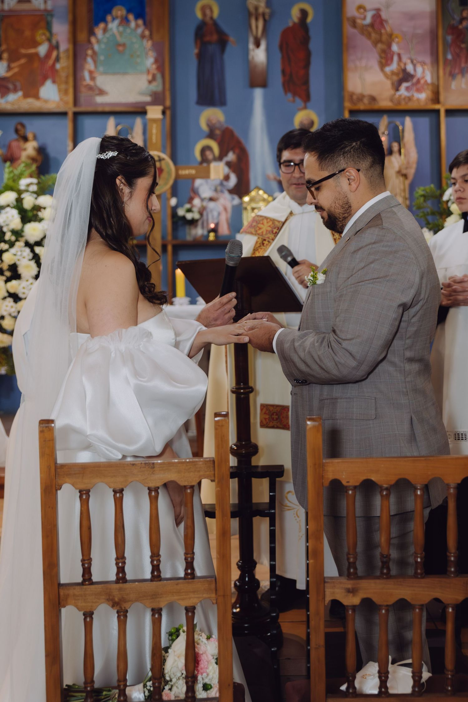 Groom places a wedding ring on the bride's finger during a Catholic church ceremony with an officiant behind them.