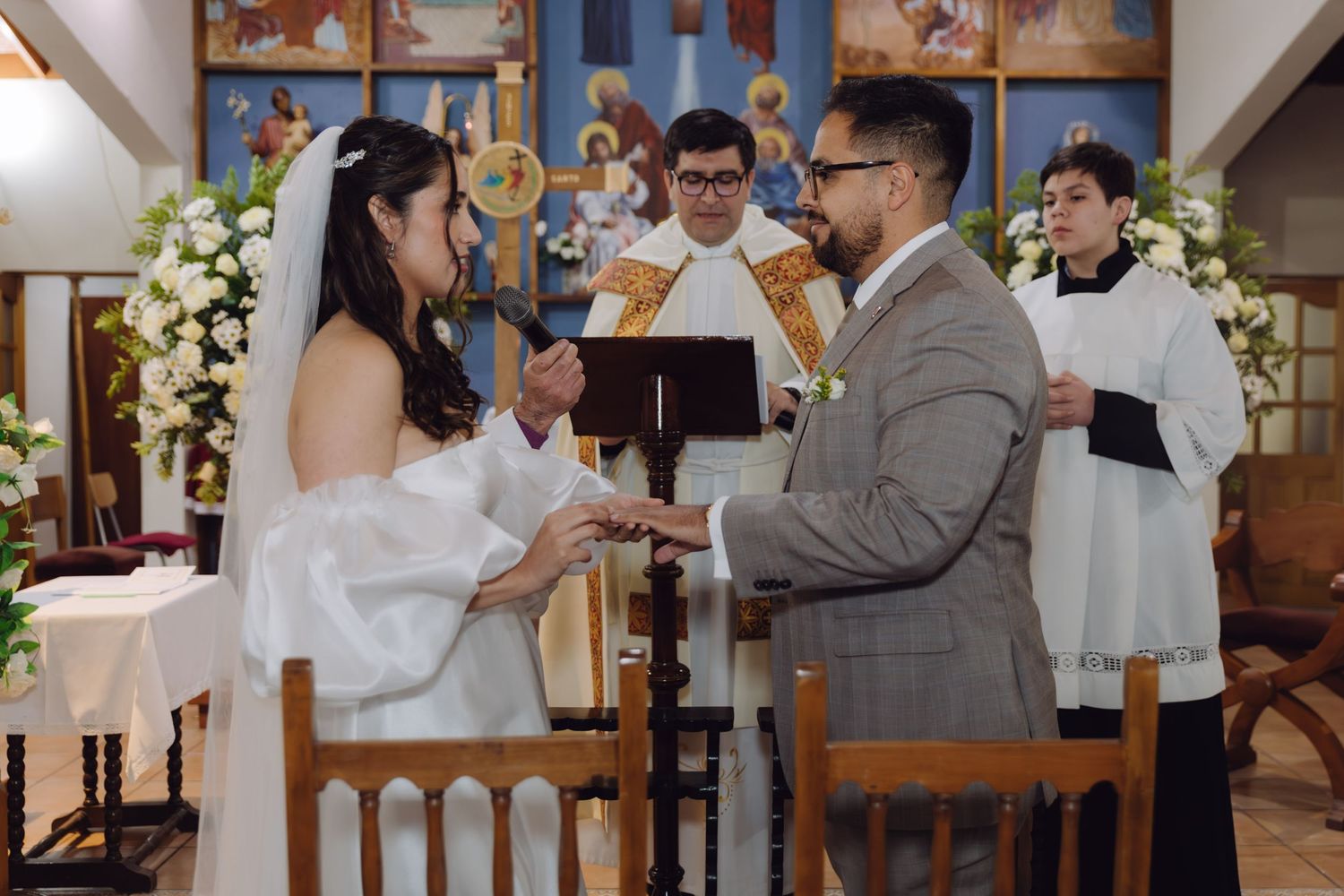 Bride and groom exchange rings at the altar of a Catholic church decorated with white floral arrangements.