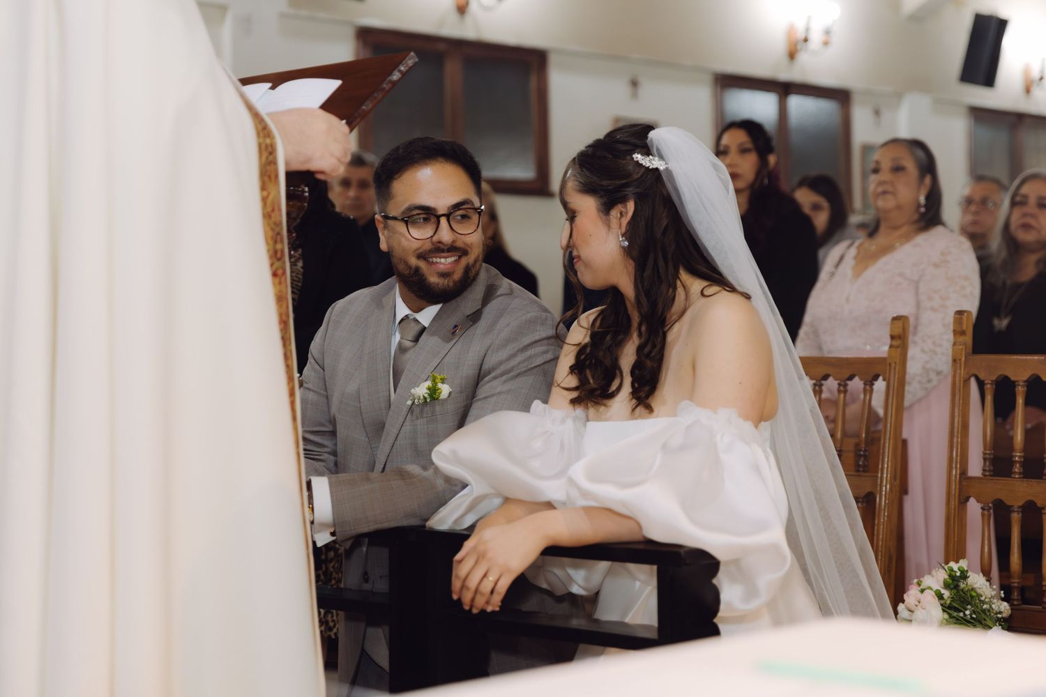 Groom smiles lovingly at the bride while seated at the church altar during their wedding ceremony.