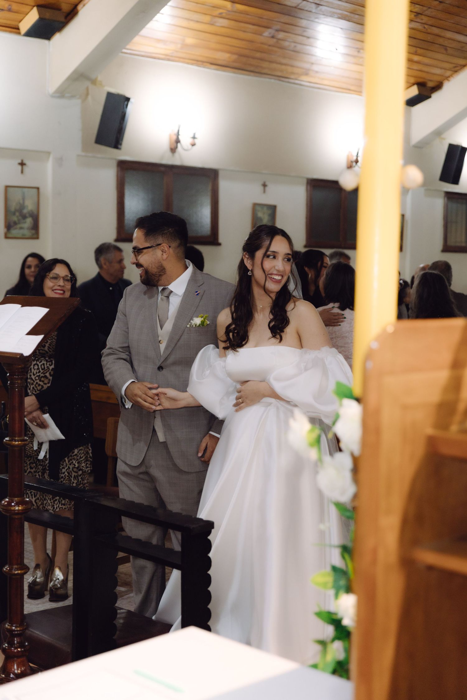 Smiling bride in white off-shoulder gown walks down church aisle with groom in gray suit during wedding ceremony.