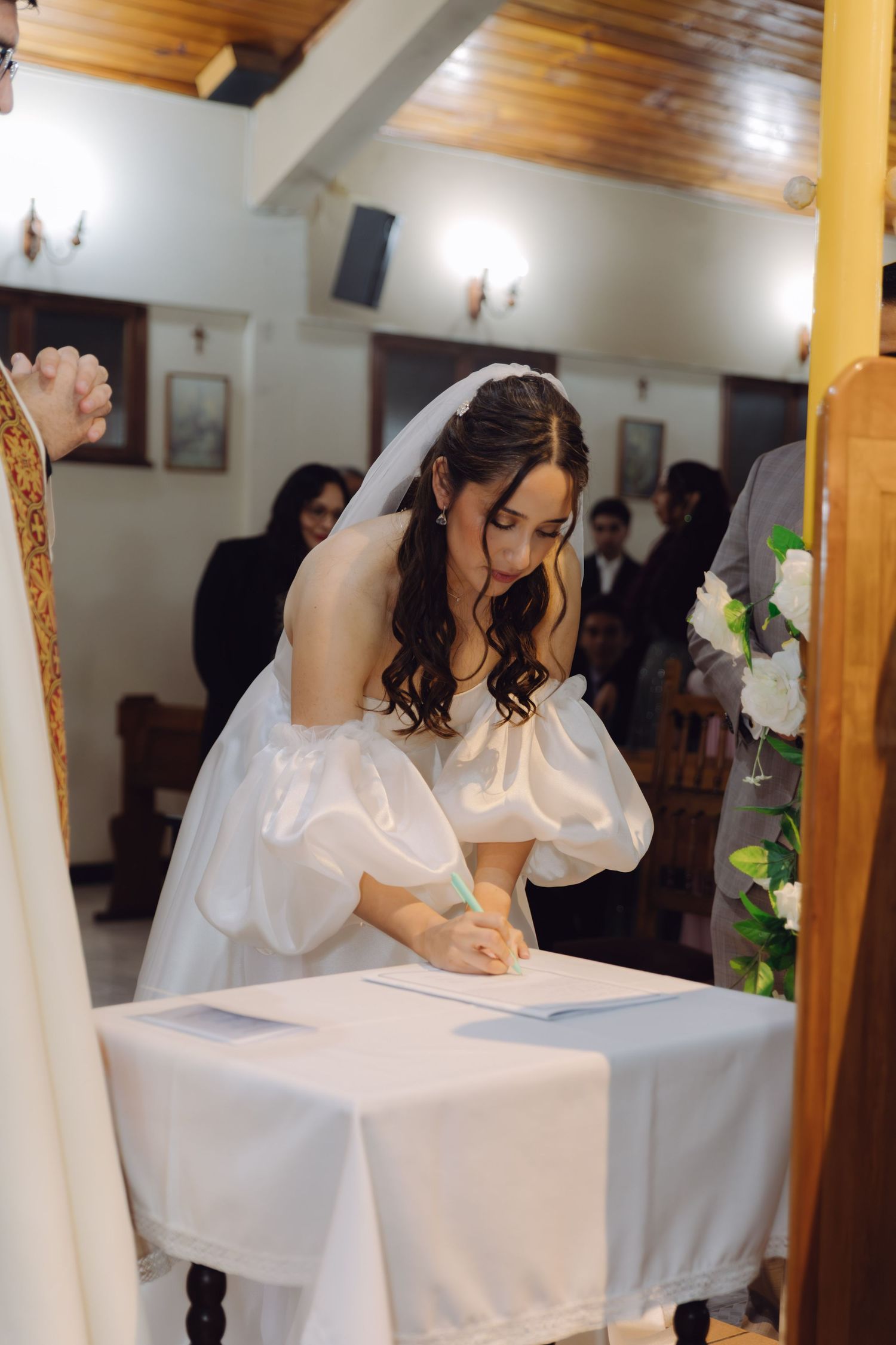 Bride in white puff-sleeve gown signs marriage documents at altar table during church wedding ceremony.