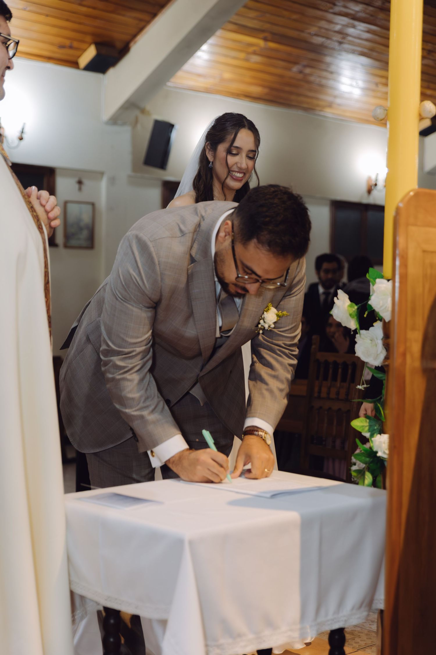 Groom in gray suit signs official marriage documents at church altar while smiling bride watches.