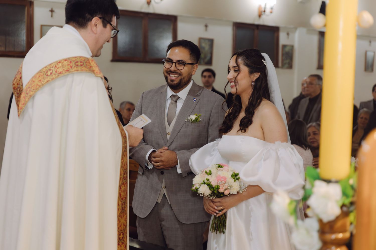 Bride holding pink bouquet and groom in gray suit exchange vows with priest during church wedding.