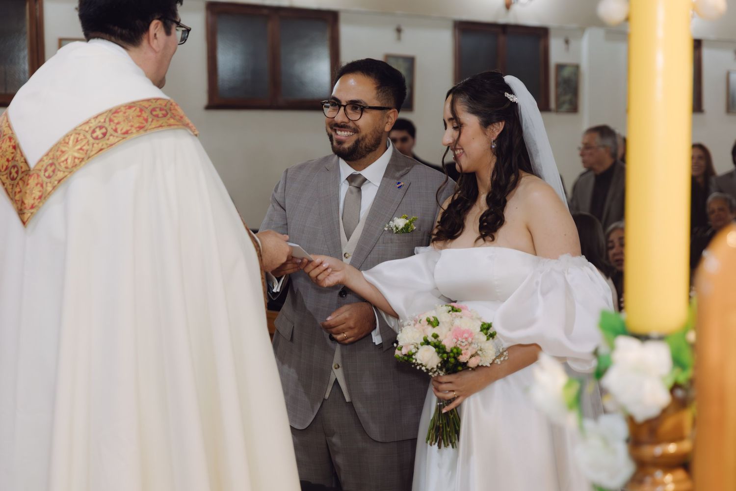 Priest officiates ring exchange ceremony as bride and groom hold hands at church altar.