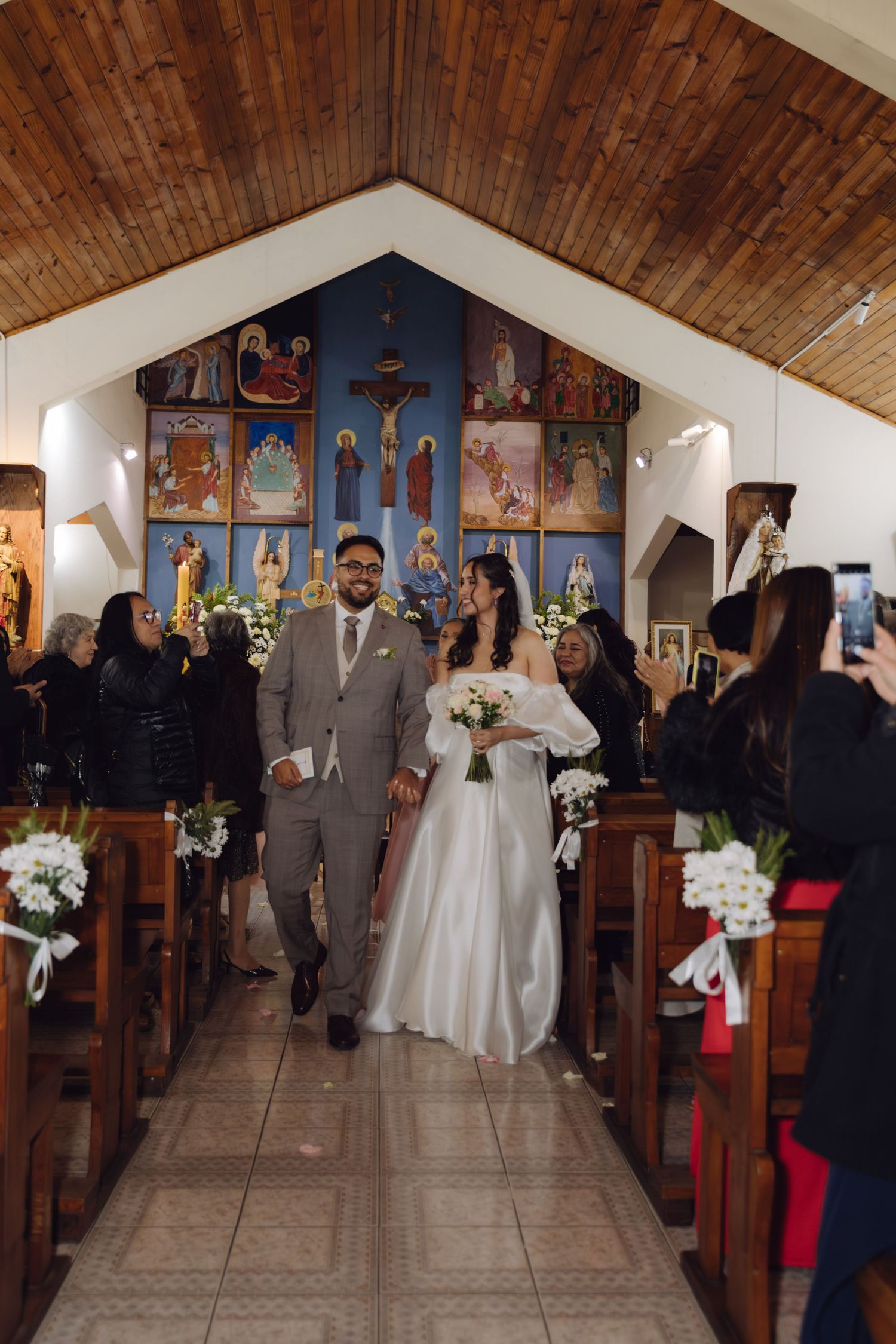 Newlyweds walk hand-in-hand down church aisle past guests after wedding ceremony with religious icons backdrop.