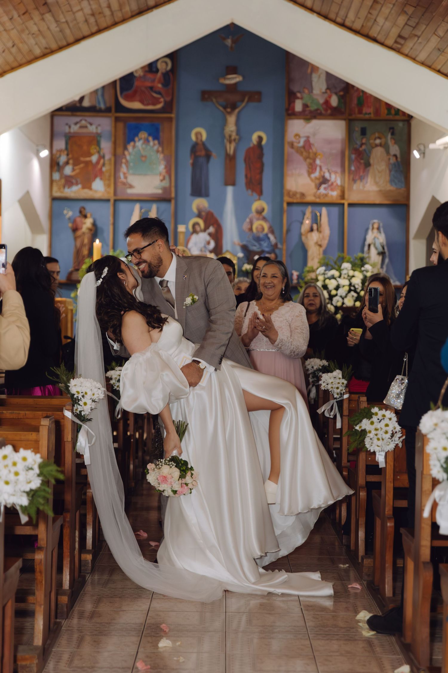 Groom lifts bride in white gown for celebratory kiss in church aisle after wedding ceremony.