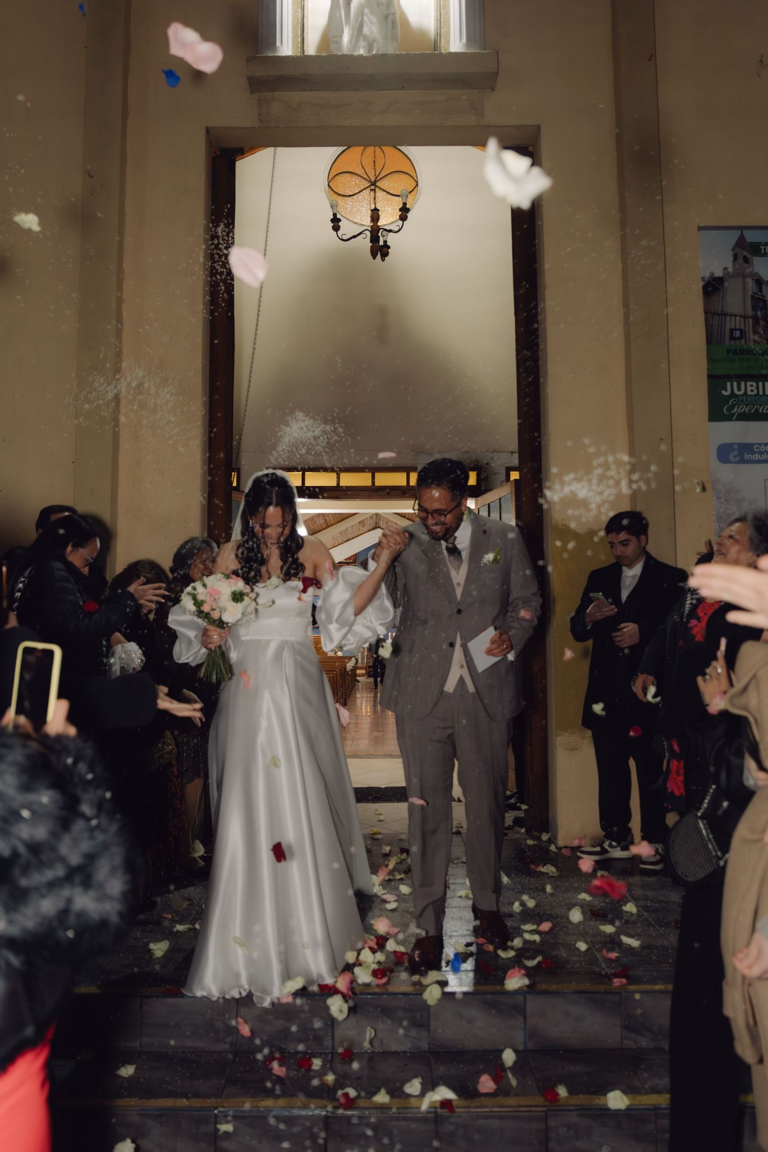Couple exits church in shower of confetti and flower petals surrounded by cheering wedding guests.