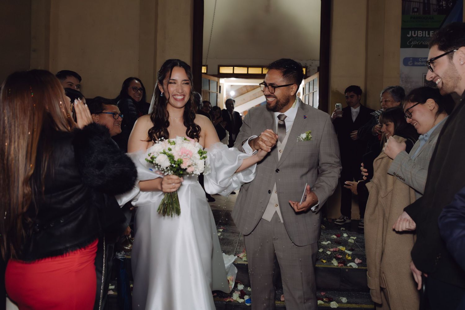 Smiling bride holding bouquet and groom in gray suit walk through cheering wedding guests.