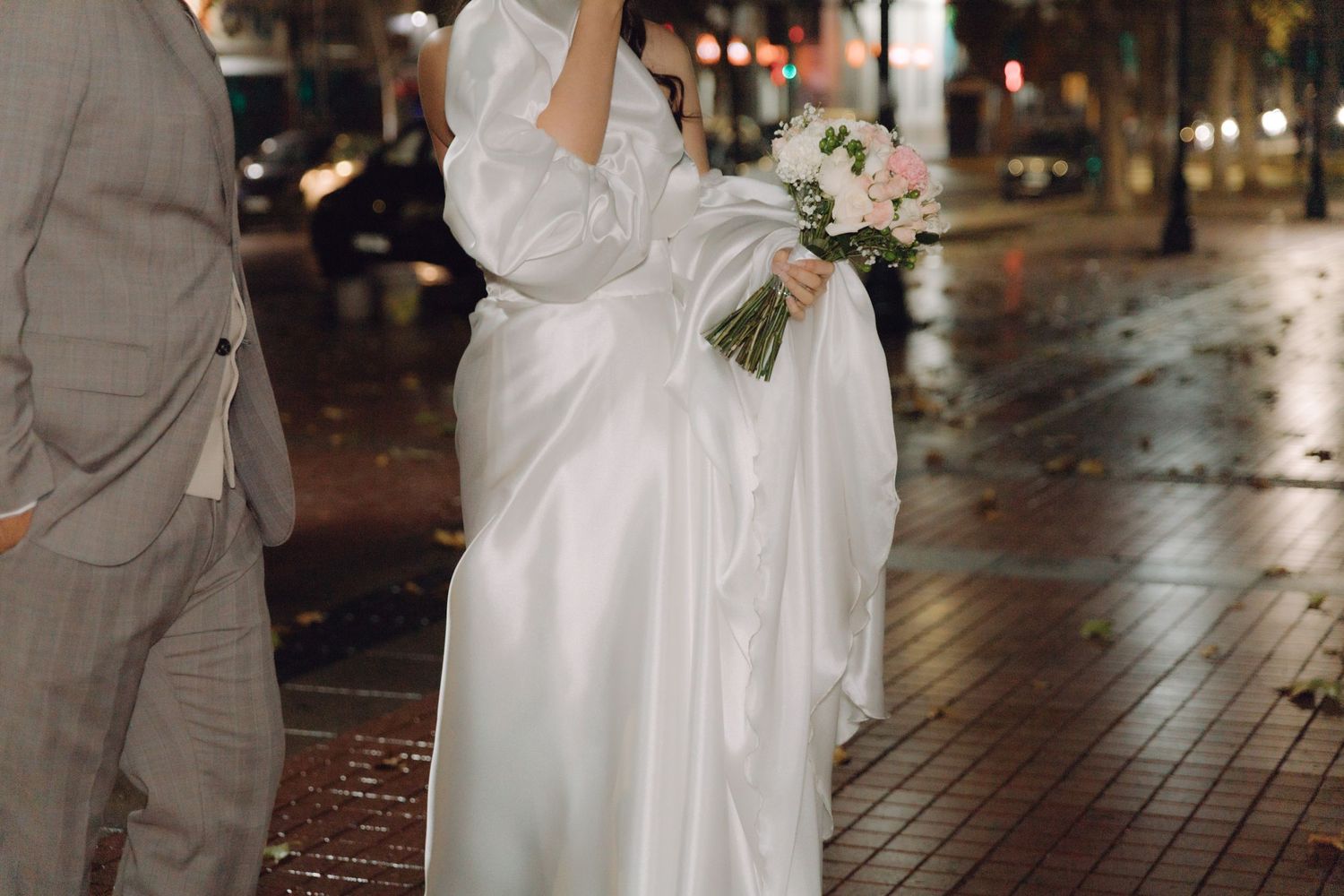 Elegant bride in white satin wedding dress holds bouquet while walking on wet city street.