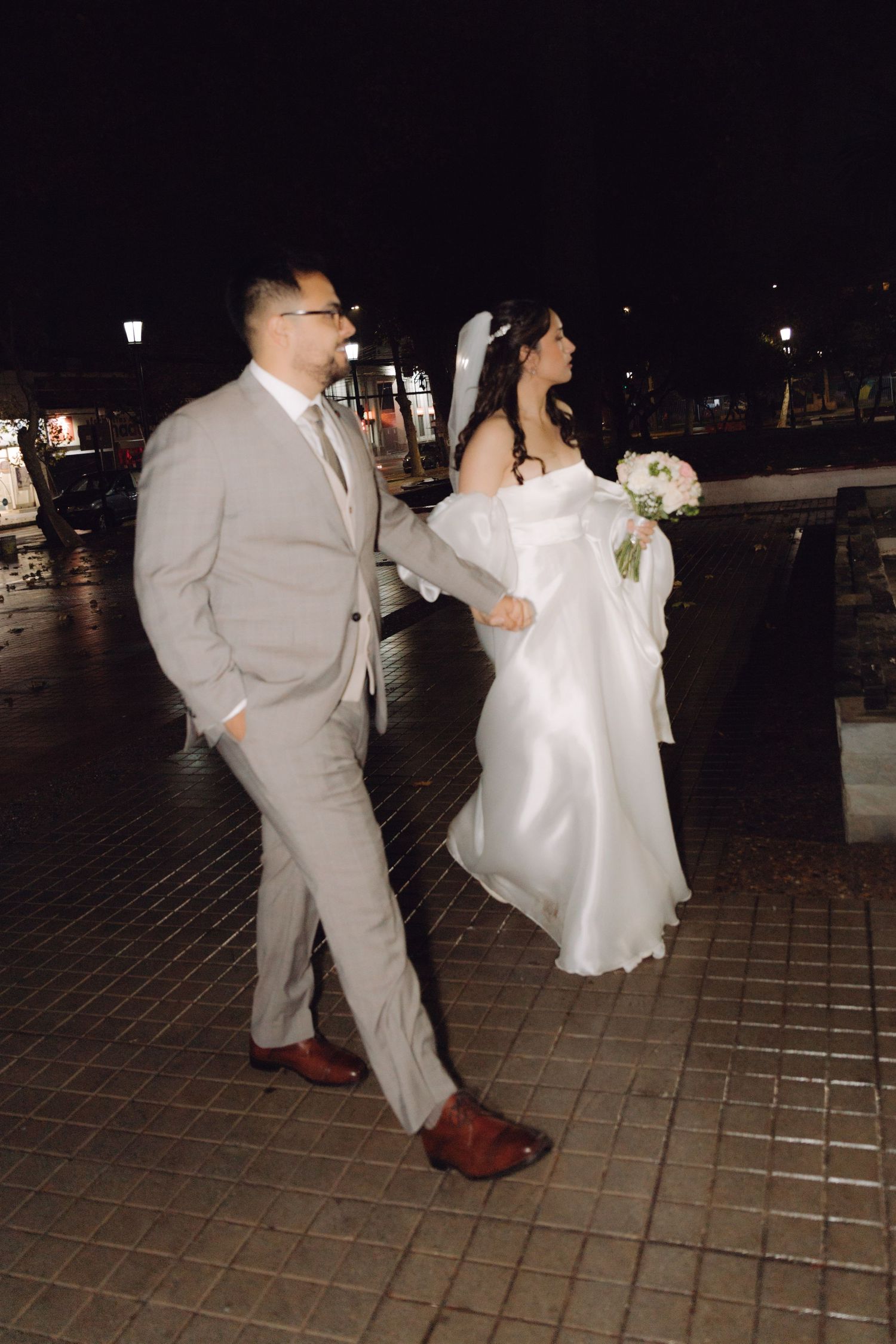 Bride in white satin gown and groom in gray suit walk together on rainy night.