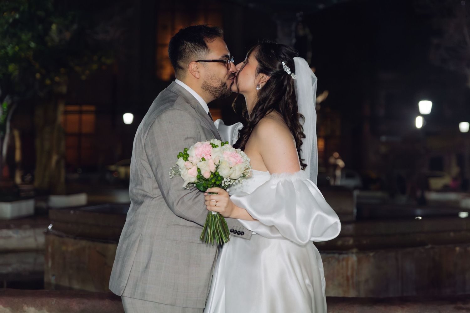 Bride and groom sharing a romantic outdoor kiss with pink bouquet at night.