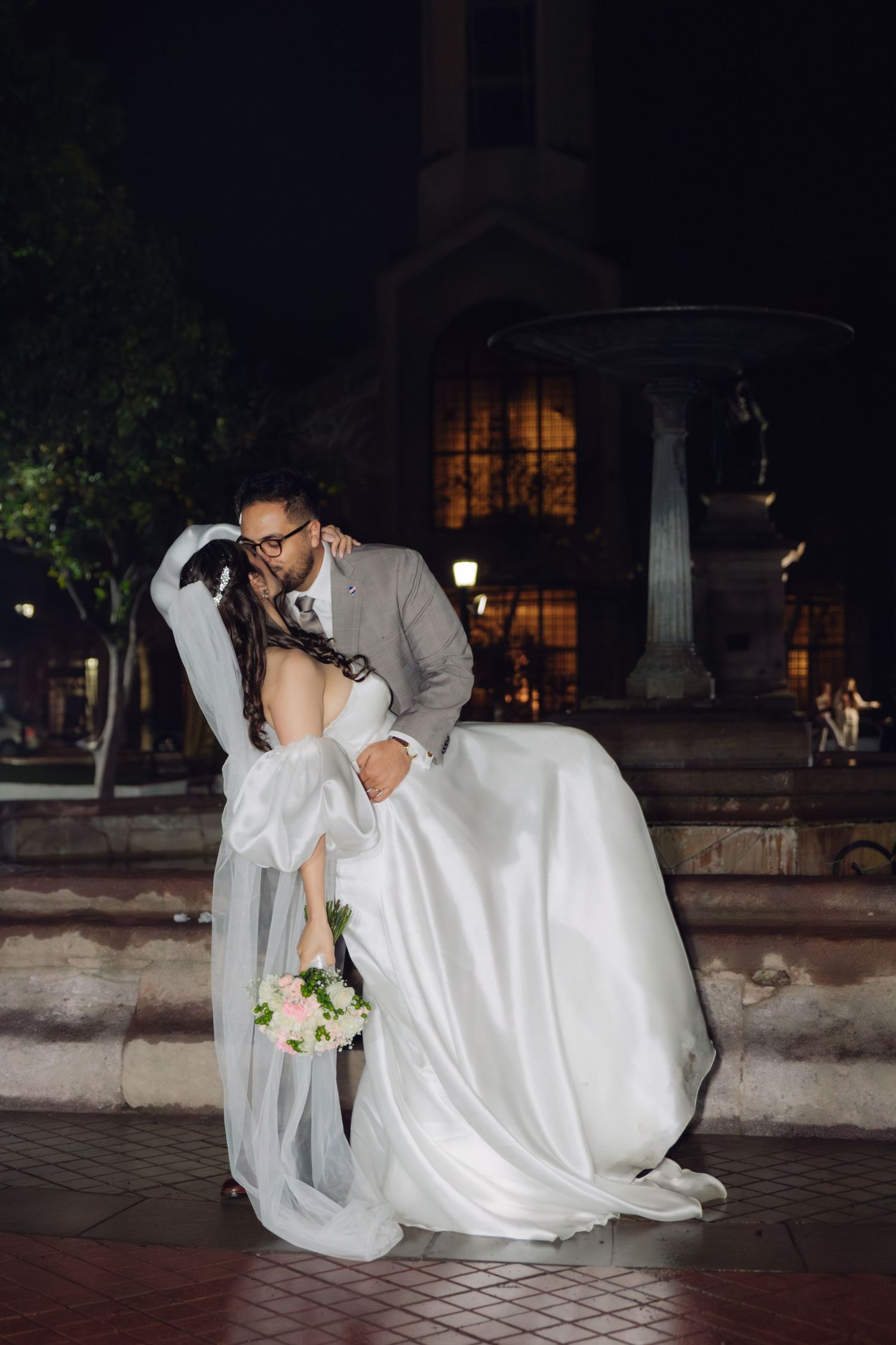 Bride and groom share a romantic dip kiss at night near a fountain, bride holding a white bouquet.