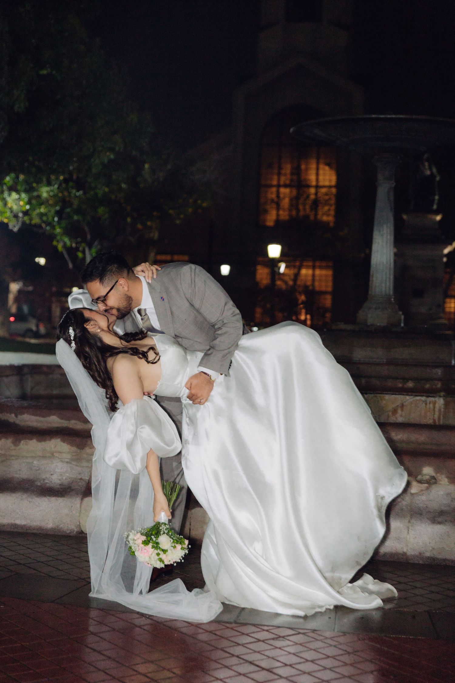 Groom dips bride for a passionate kiss outdoors at night near an illuminated fountain.