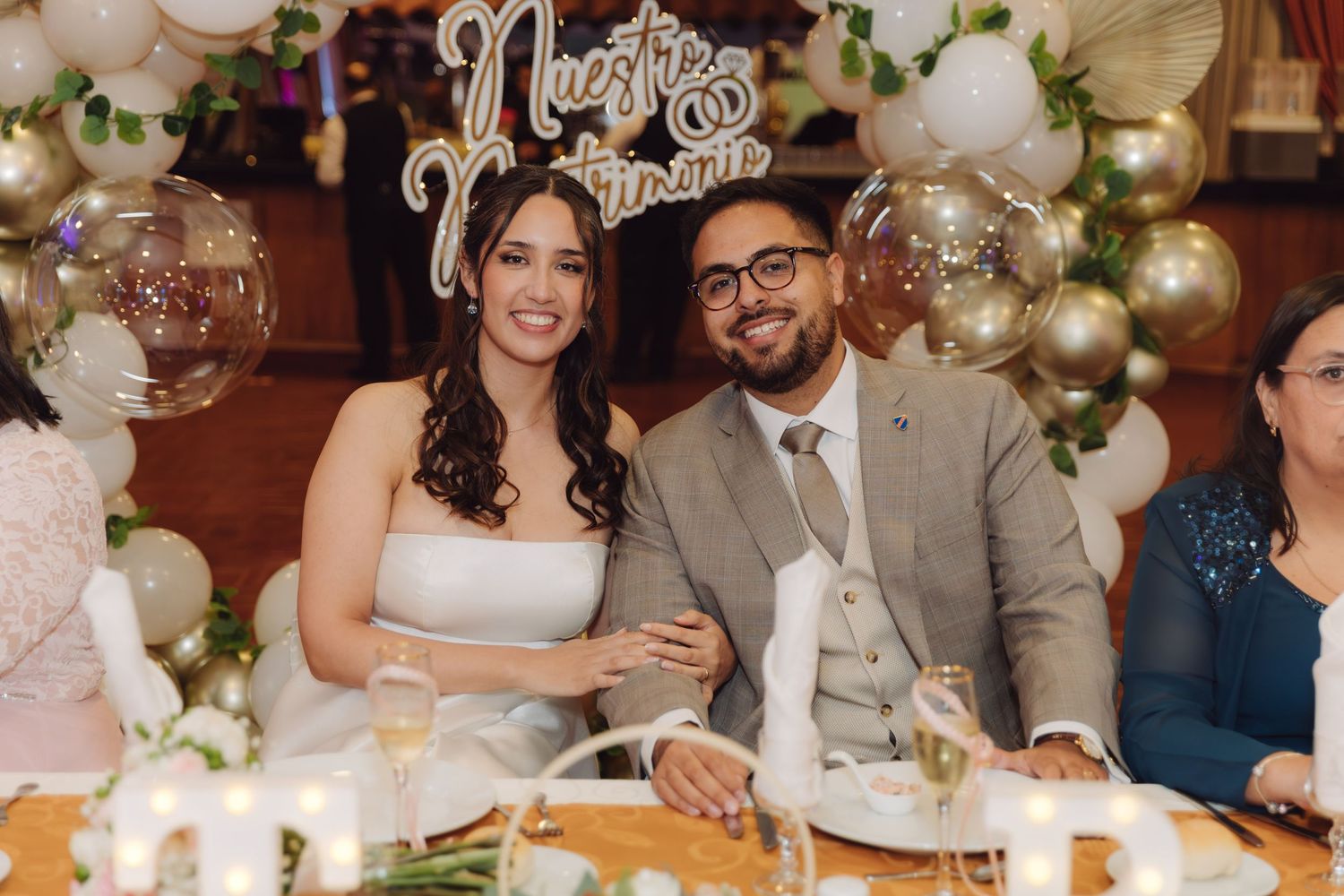 Smiling bride and groom sit together at their wedding reception table with champagne glasses.