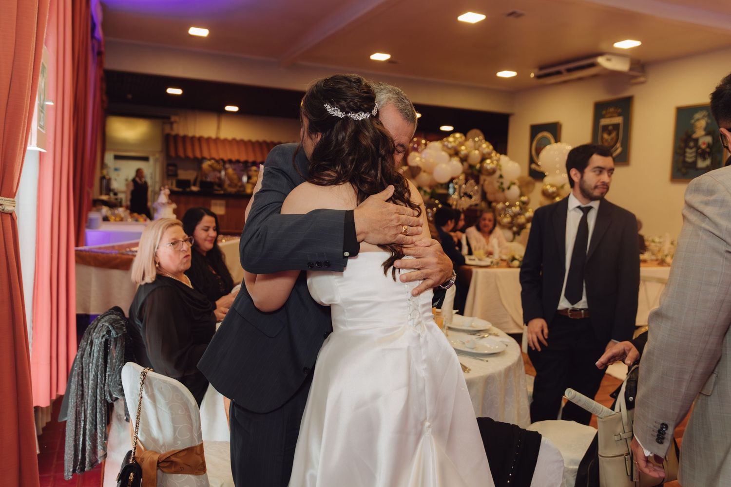 Bride in white gown shares a heartfelt hug with an older guest at the wedding reception.
