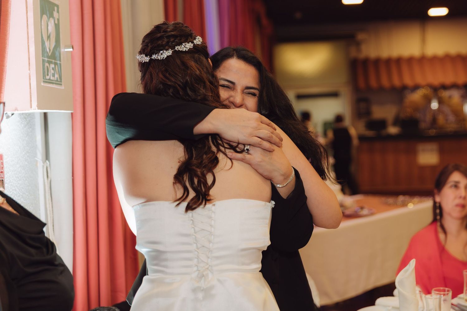 Bride in strapless white dress receives a warm embrace from a smiling female guest at the reception.