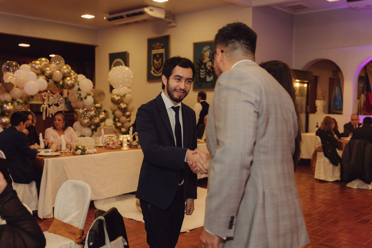 Two well-dressed male wedding guests share a handshake at an elegant balloon-decorated reception.