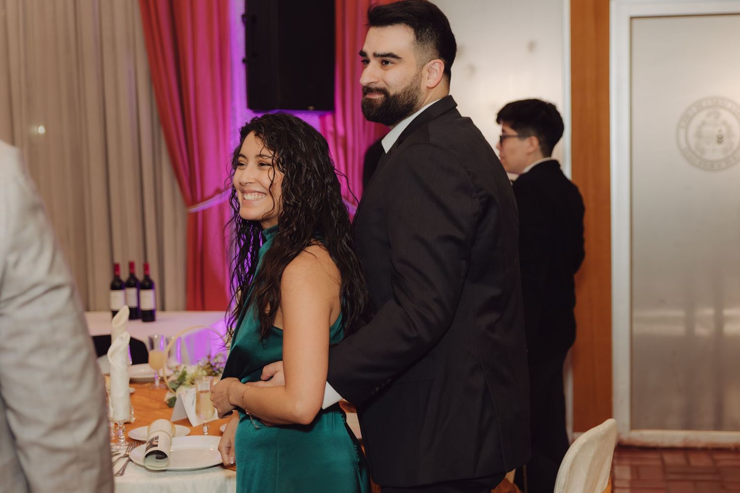 Smiling woman in green dress stands with bearded man in black suit at elegant wedding reception.