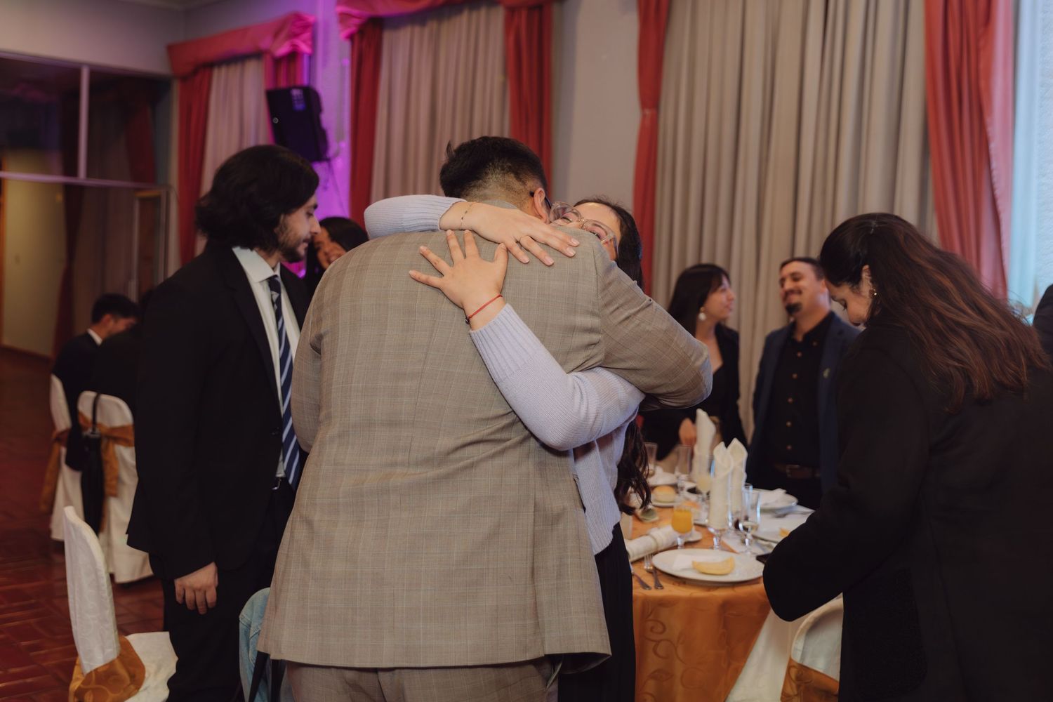 Guests embrace warmly at a wedding reception in an elegant banquet hall.