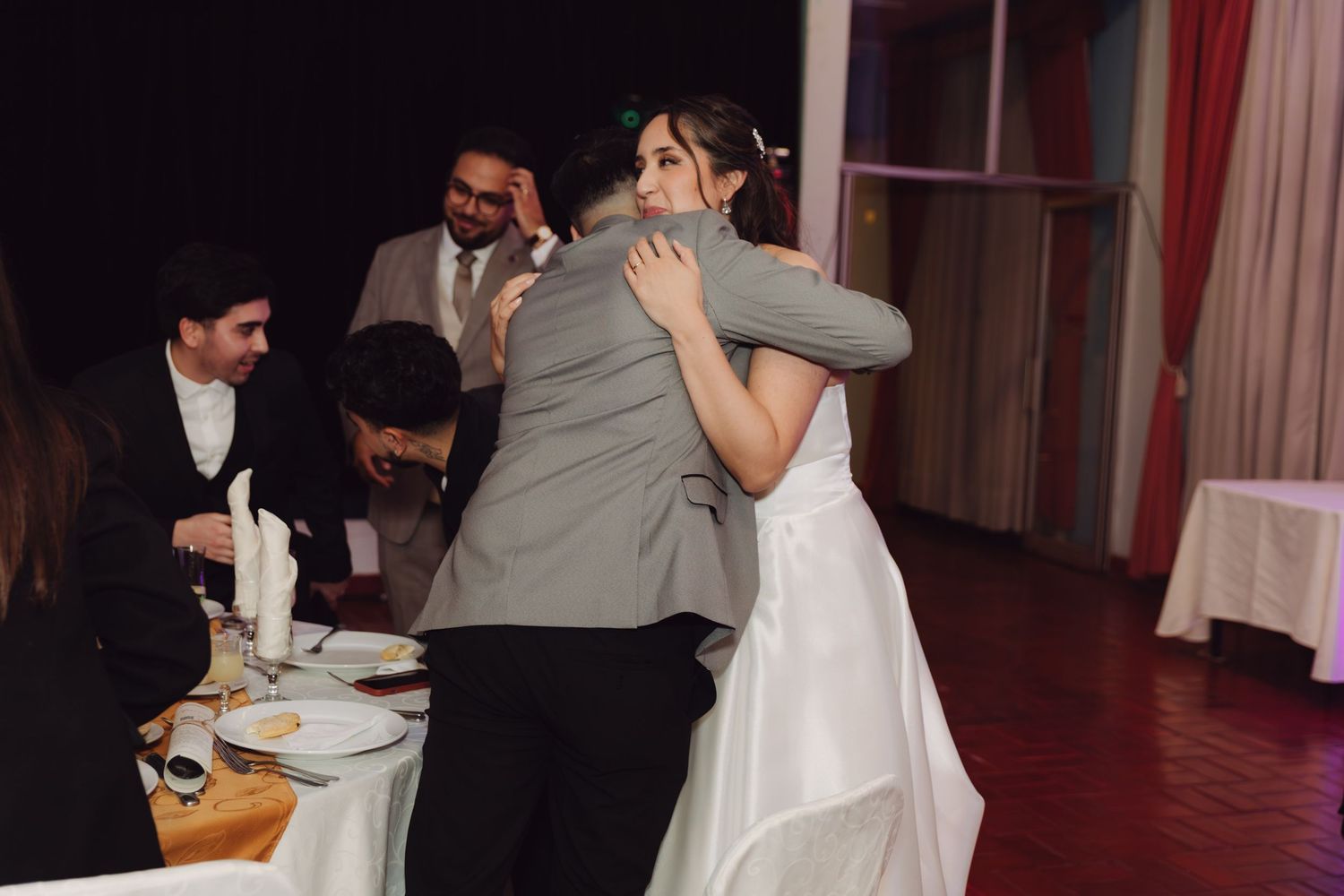 Bride in white wedding dress shares warm embrace with guest at reception.
